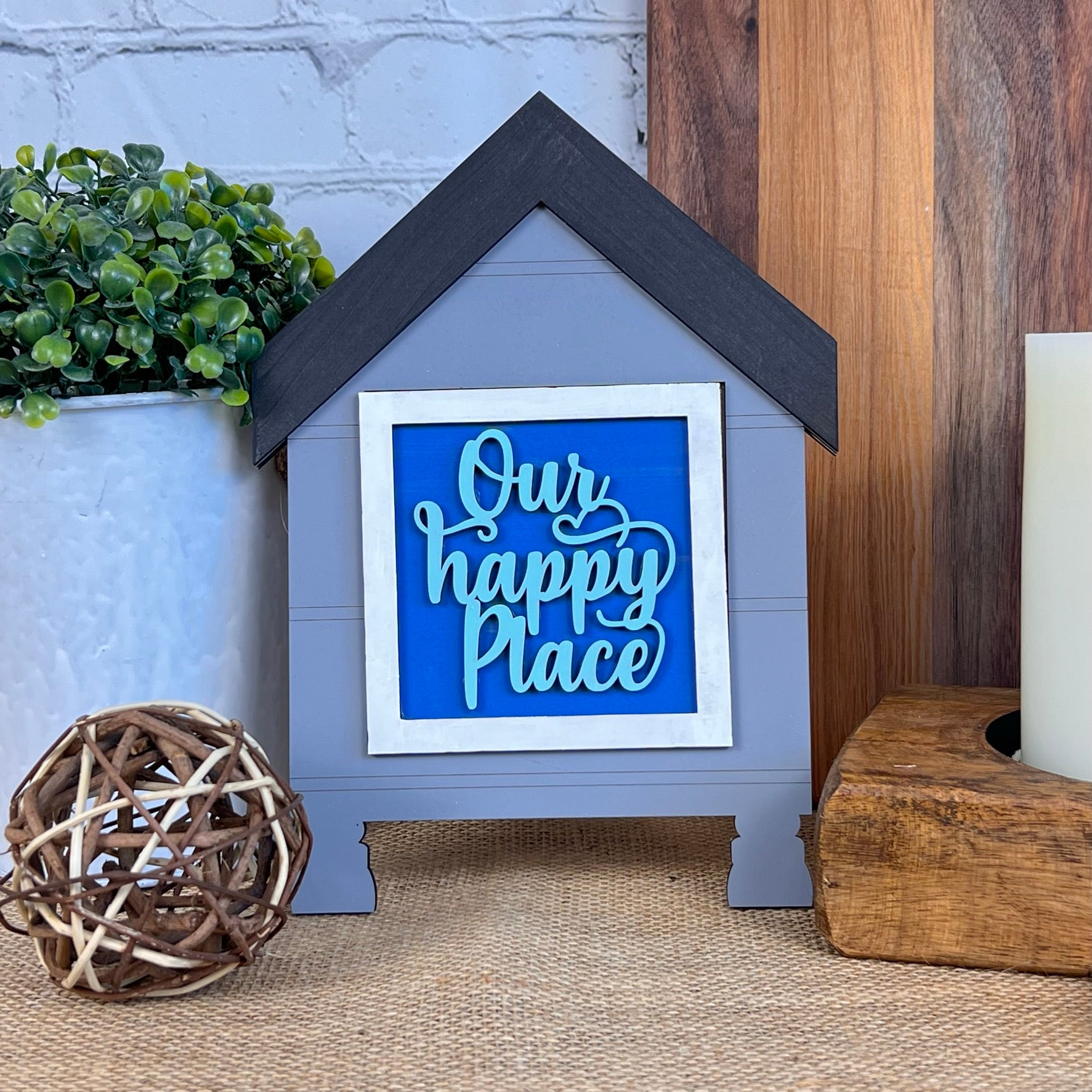 The Home Family Interchangeable Squares by Janet's Craft Corner is a charming small house-shaped decor item that includes an interchangeable blue sign displaying the message "Our happy place." Ideal for versatile home decor enthusiasts, it rests on a wooden surface beside a plant, wicker ball, and candle holder, with a brick wall as the backdrop.