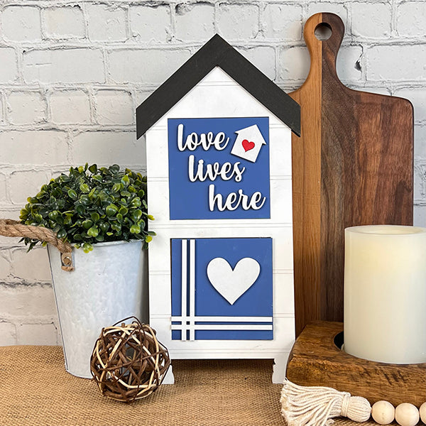 The Home Family Interchangeable Squares by Janet's Craft Corner features a charming house-shaped plaque adorned with the words "love lives here" and a heart symbol. It's set amidst a potted plant, wooden cutting boards, and mini signs, creating cozy home decor against a white brick wall that adds charm to any space.