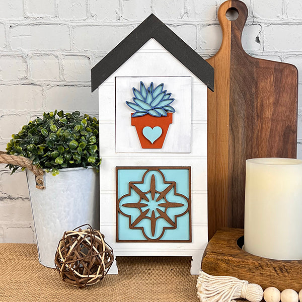 A decorative arrangement featuring the Home Family Interchangeable Squares from Janet's Craft Corner includes a small wooden house adorned with succulent and geometric designs, complemented by a potted plant, wooden cutting boards, a candle, and a wicker ball against a brick wall backdrop. This charming setup effortlessly enhances any space with its rustic appeal.