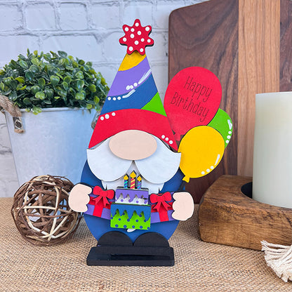 A vibrant Birthday Gnome from Janet's Craft Corner holds a cake and balloons, one of which is inscribed with "Happy Birthday." It features a hat topped with a star. Perfect as home décor, it stands on a textured surface surrounded by plants, candles, and charming wall decorations.