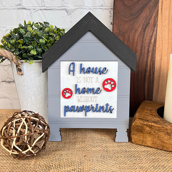 Janet's Craft Corner presents the Dog Interchangeable Squares, a delightful decorative piece shaped like a house with the phrase, "A house is not a home without pawprints," accented by two red pawprint icons. Ideal for incorporating farmhouse elements, it pairs beautifully with a potted plant, wicker ball, and candles to enhance a cozy ambiance on any table.