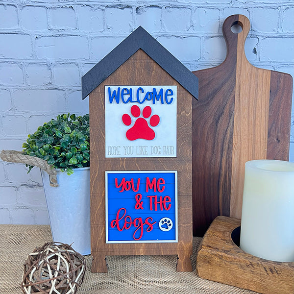 Janet's Craft Corner presents the Dog Interchangeable Squares, a decorative wooden house-shaped sign adorned with "WELCOME, Hope You Like Dog Hair" and "You Me & The Dogs," complete with an adorable paw print design. Surrounded by farmhouse accents such as a small plant, candle, and cutting boards against a brick wall backdrop, this piece beautifully captures rustic charm.