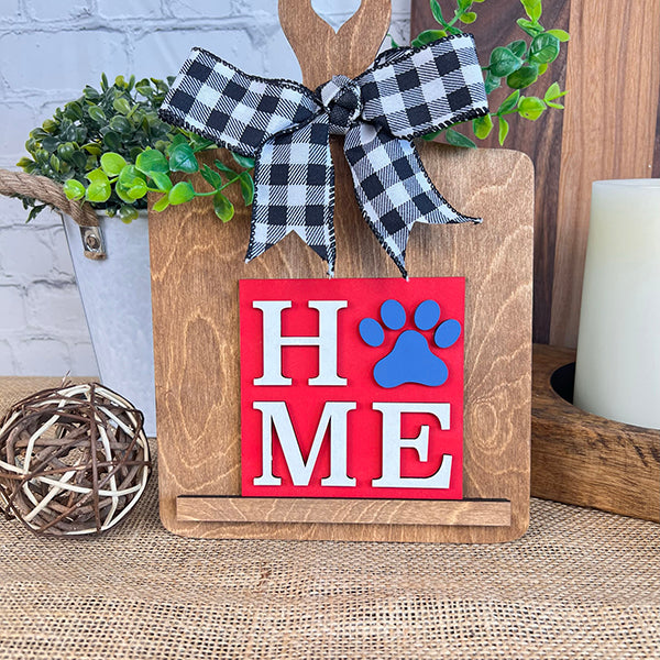 Introducing the Dog Interchangeable Squares from Janet's Craft Corner: This charming wooden board features a black-and-white checkered bow and spells out "HOME" with a delightful blue paw print replacing the "O." Ideal for tiered trays, it pairs beautifully with greenery, a woven sphere, and a candle on a rustic surface to add cozy farmhouse elements.