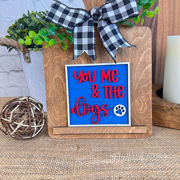 The Dog Interchangeable Squares by Janet's Craft Corner feature a charming wooden sign with the phrase "You Me & the Dogs" in red on a blue background, enhanced with a paw print. This decorative piece exudes farmhouse charm as it sits on a wooden stand adorned with a black and white checkered bow, beautifully displayed among greenery and decorative tiered trays.