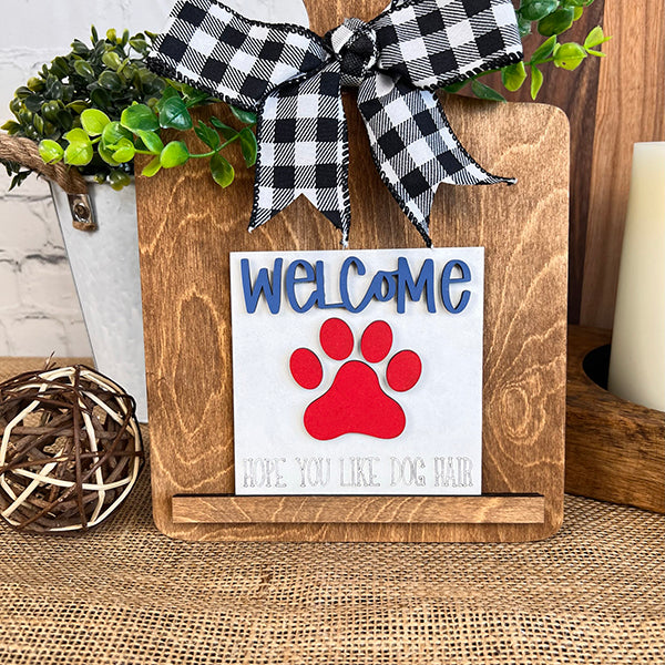 A wooden stand from Janet's Craft Corner showcases rustic farmhouse appeal with a sign adorned with a checkered ribbon from the Dog Interchangeable Squares collection. The sign features the word "Welcome" alongside a red paw print, with "Hope you like dog hair" written beneath it. Surrounding tiered trays display items like a plant, wicker ball, and candle, creating a cozy homage to our love for dogs.