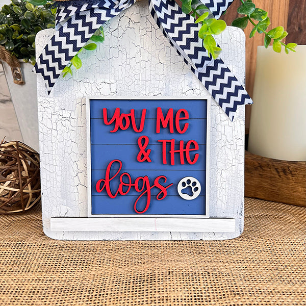 A decorative piece from Janet's Craft Corner, the Dog Interchangeable Squares features a blue background with red text that reads "You Me & the Dogs" alongside a paw print. The sign is framed in rustic white wood and embellished with a black-and-white zigzag ribbon on burlap cloth, making it an ideal addition to tiered trays for that farmhouse touch.