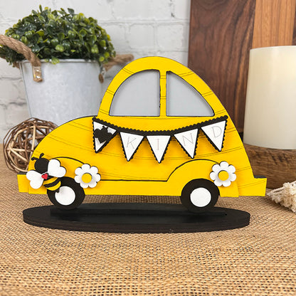 A decorative wooden car from Janet's Craft Corner, featuring the Bee Volkswagen design in yellow, comes with a "KIND" banner and floral accents showcased on a burlap mat. In true farmhouse decor style, it is complemented by a plant, candle, and decorative ball adorning the wooden surface in the background.