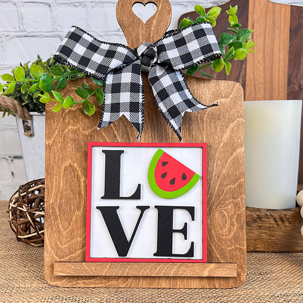 Introducing the Watermelon Interchangeable Squares by Janet's Craft Corner, a delightful farmhouse decor piece featuring the word "LOVE," with a watermelon slice creatively substituting for the "O." This charming item is accented with a black and white checkered bow, greenery, a candle, and a woven ball. It's an ideal choice for enhancing tiered trays against textured backdrops.