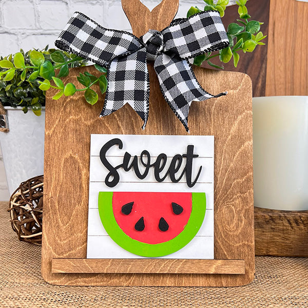 Janet's Craft Corner's Watermelon Interchangeable Squares, adorned with a black and white checkered bow on top and featuring the word "Sweet" alongside a vibrant watermelon slice, are the perfect addition to farmhouse decor. When placed among tiered trays on a table, they are beautifully accented by greenery and a white candle in the background.