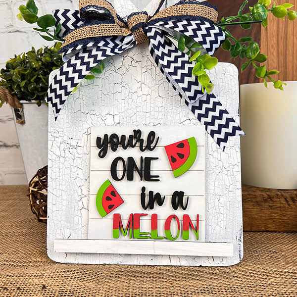 The Watermelon Interchangeable Squares by Janet's Craft Corner feature a charming decorative sign with the phrase "you're one in a melon" in vibrant letters, adorned with watermelon slice designs. This piece is anchored by a chevron-patterned ribbon on a rustic white board, making it perfect for tiered trays and adding charm to farmhouse decor, especially when displayed alongside leafy plants.