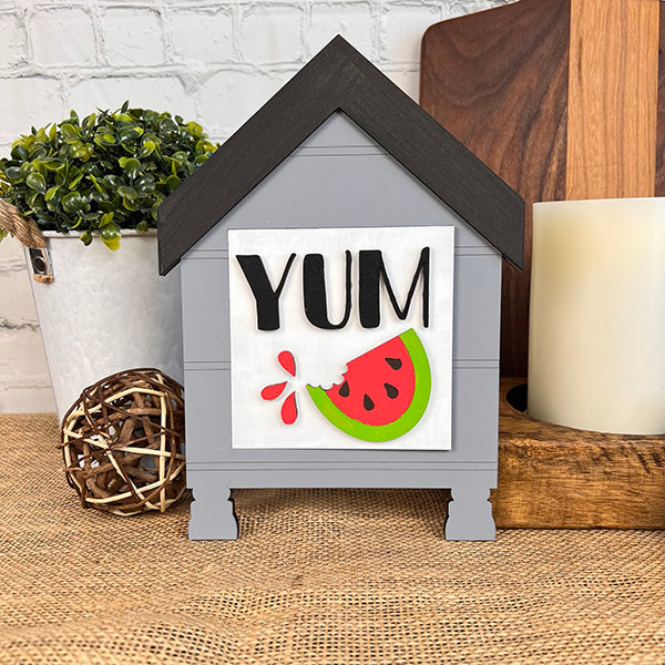 Introducing the Watermelon Interchangeable Squares from Janet's Craft Corner: A delightful decorative sign shaped like a gray house with a black roof, perfect for enhancing your farmhouse decor. This charming piece displays the word "YUM" in black along with a vibrant illustration of a watermelon slice. Ideal for tiered trays, it pairs beautifully when placed on burlap surrounded by plants and complemented by a large candle.