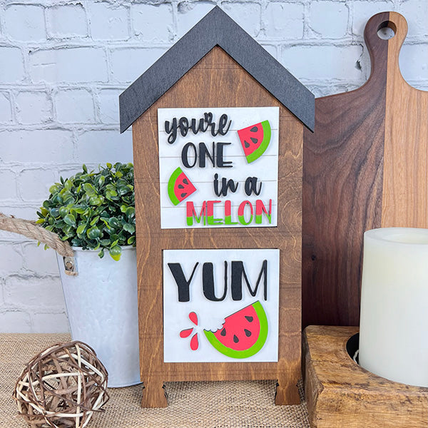 The Watermelon Interchangeable Squares from Janet's Craft Corner showcase a decorative wooden design in the shape of a house. This charming piece includes the phrases "You're one in a melon" and "Yum," adorned with delightful watermelon illustrations that enhance its rustic appeal. Perfect for farmhouse decor, it adds personality to tiered trays or any tabletop setting.