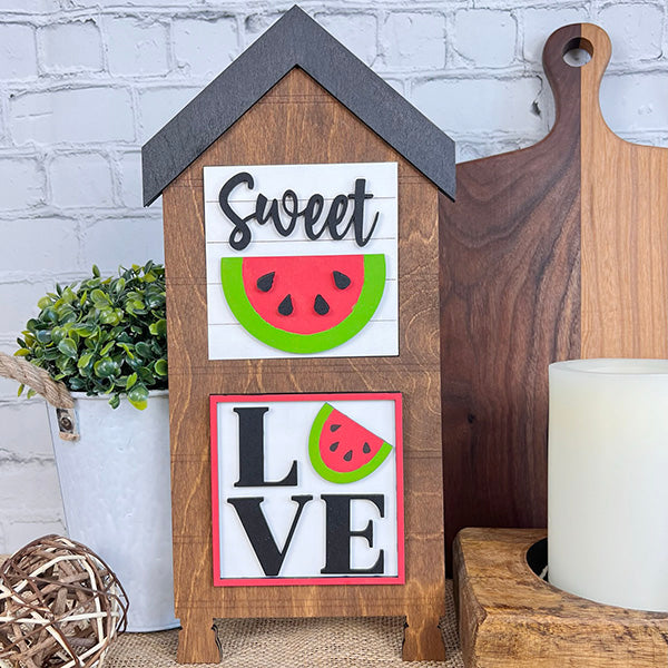 A decorative piece from Janet's Craft Corner, the Watermelon Interchangeable Squares, is shaped like a house and adorned with "Sweet" and "LOVE" words alongside charming watermelon designs. Ideal for tiered trays or enhancing your farmhouse decor, it elegantly rests against a white brick wall, complemented by a plant, cutting boards, and a candle.