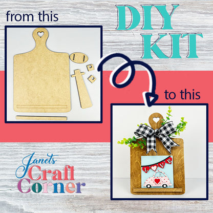 Introducing Janet's Craft Corner's Breadboard Interchangeable Squares Holder DIY kit, which transforms a plain wooden cutout into an elegantly decorated sign holder featuring a heart handle. The completed design includes a charming small car, a "LOVE" heart, lush greenery, and a chic black-and-white bow—ideal for displaying interchangeable square signs.