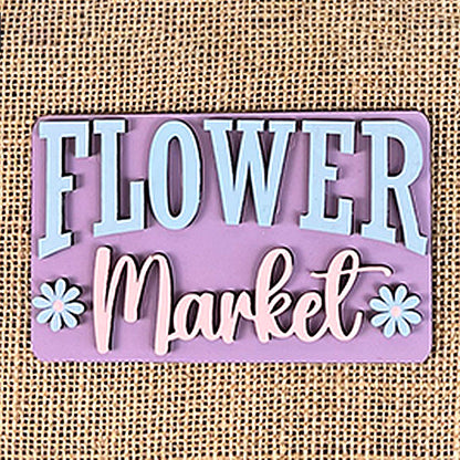 A decorative sign featuring "Flower Market" in light blue and white letters graces a pink background, adorned with small daisy illustrations on a textured burlap surface. This charming piece, reminiscent of a hand-painted style, pairs beautifully with the Flower Market Mini Shelf Sitters from Janet's Craft Corner.