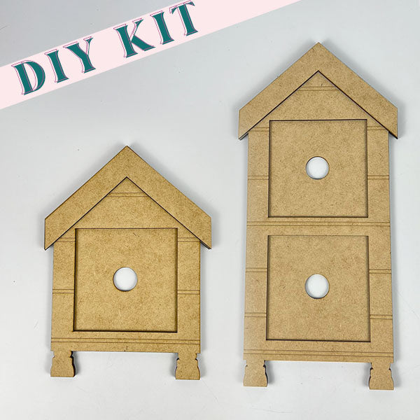 This delightful farmhouse décor DIY kit, "House Interchangeable Squares Holders" by Janet's Craft Corner, features two MDF birdhouses. The petite house includes a single circular entrance, while the larger one is designed with two entrances beneath pointed roofs. Ideal for your seasonal craft kit collection, the label "DIY KIT" is clearly highlighted in the top left corner.