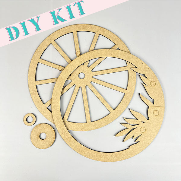 The Interchangeable Wagon Wheel Door/Wall Hanger from Janet's Craft Corner features wooden cutouts perfect for home décor. It includes a circular wheel, a decorative ring with leaf patterns, and two small circular pieces, making it ideal for creating a unique door hanger against a light background.