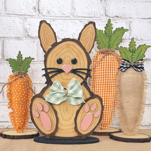 The Wood Slice Bunny Shelf Sitter by Janet's Craft Corner is an enchanting Easter decoration featuring a wooden bunny adorned with a green checkered bow tie, accompanied by three carrot replicas crafted from orange and burlap fabric. This charming scene is perfectly set against a textured white brick wall, providing a rustic accent to your festive decor.