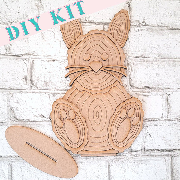 A Wood Slice Bunny Shelf Sitter by Janet's Craft Corner is displayed on a white brick wall, making it an ideal decoration for Easter. The bunny shows layered details of its face, ears, and paws. An oval piece accompanies it. The words "DIY KIT" appear in the top left corner.